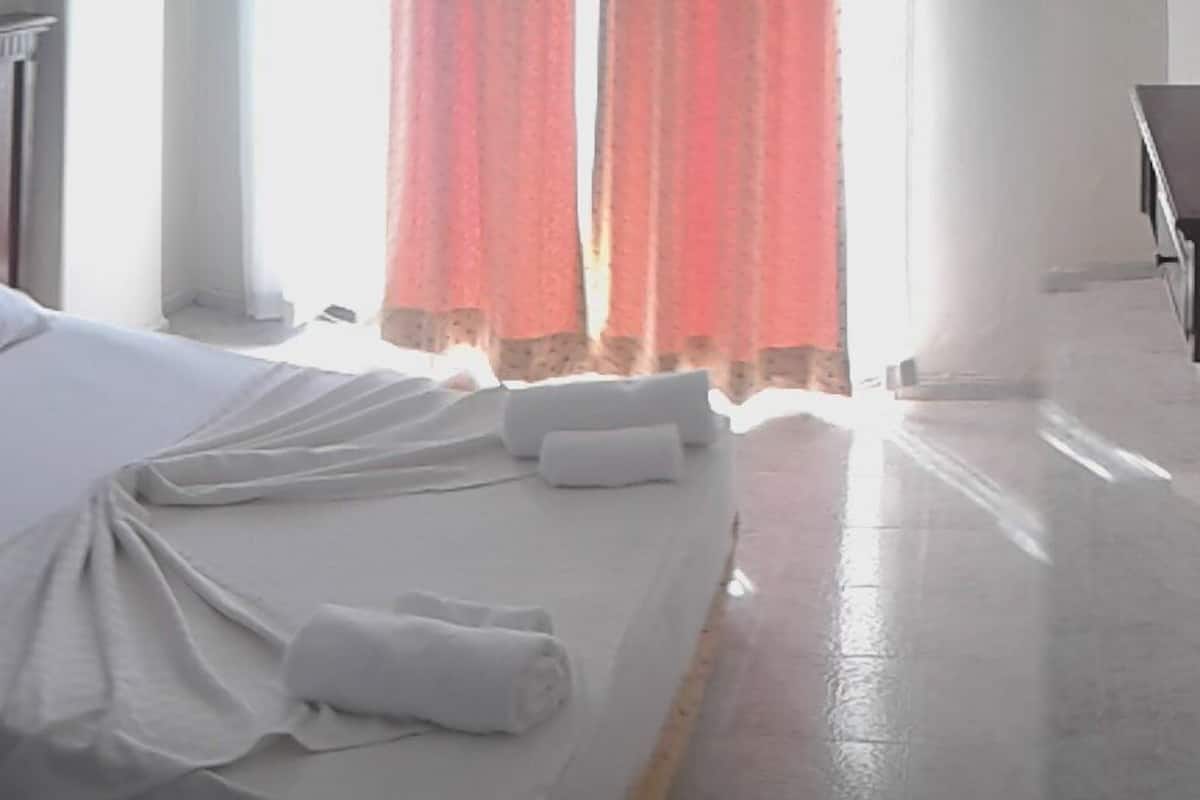 Una cama bien hecha con sábanas blancas y un cabecero de madera, una mesita de noche pequeña y una ventana con cortinas rojas.