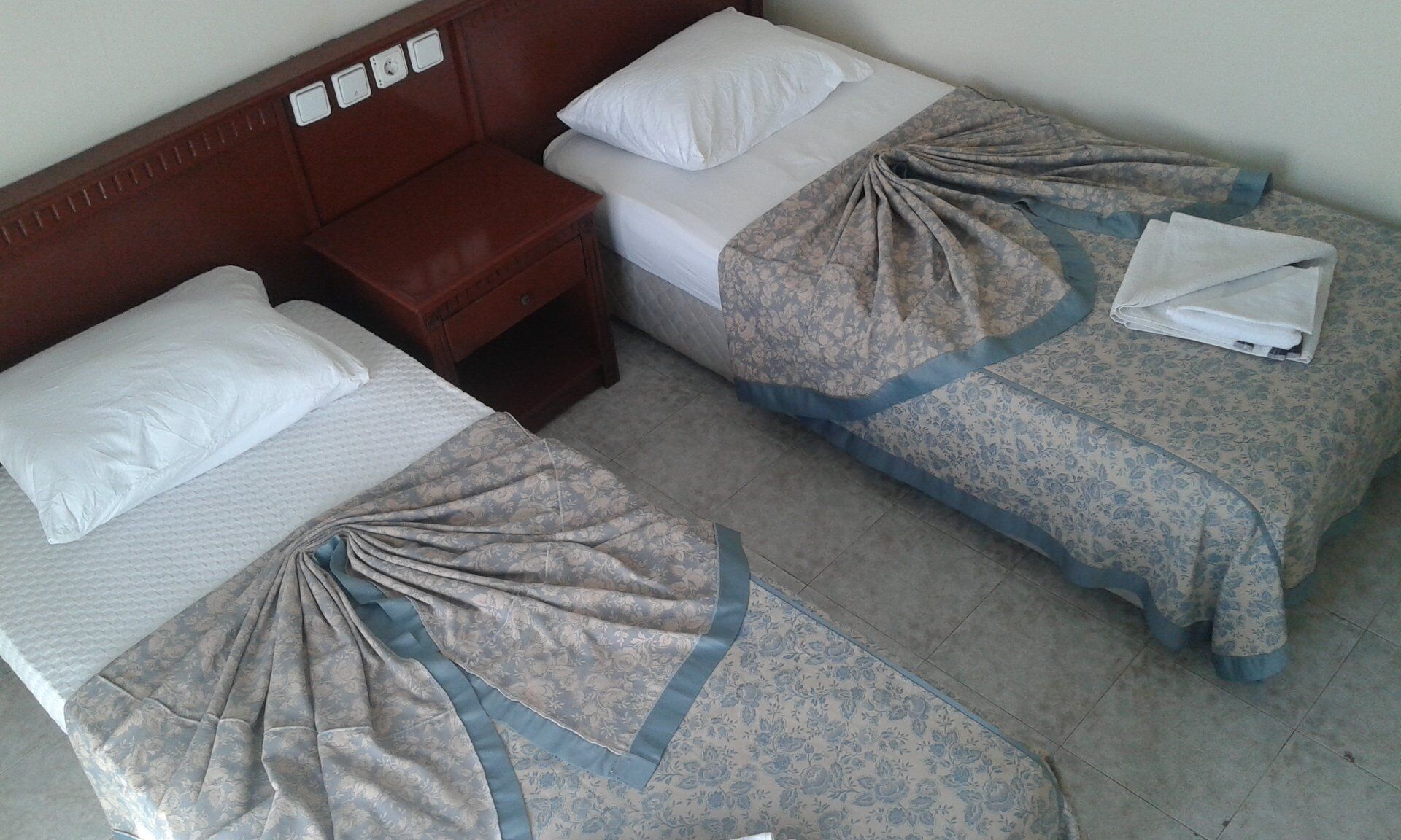 A hotel room with two single beds, each with a pillow and a blanket. There is a wooden nightstand with a lamp between the beds.