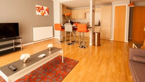 A modern living room with a TV, a coffee table, and a red patterned rug. There are orange chairs and a kitchen in the background. - ByEvo 1 Brabloch GLA Apartment  (Paisley)