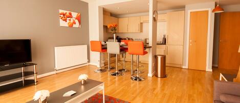 A modern living room with a TV, a coffee table, and a red patterned rug. There are orange chairs and a kitchen in the background.
