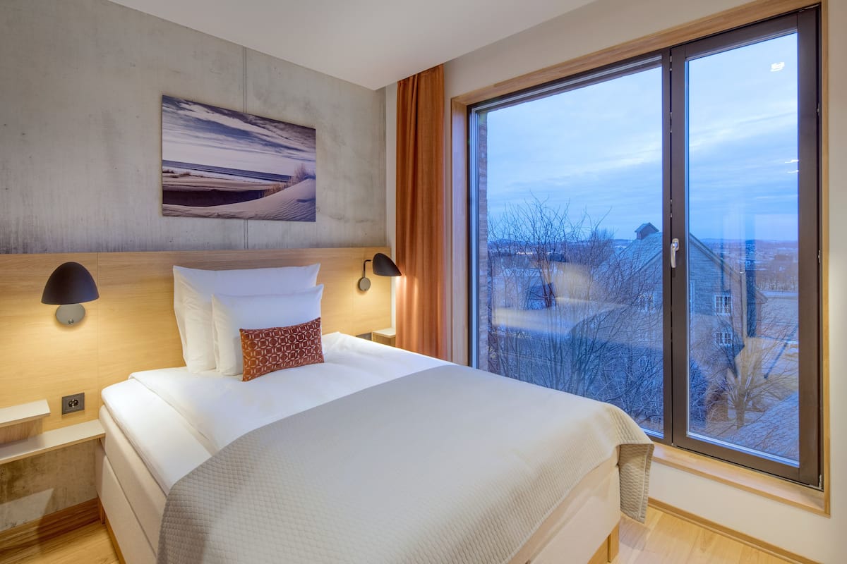 A modern bedroom with a large bed, a nightstand, a wall-mounted lamp, and a large window with a view of the outside.