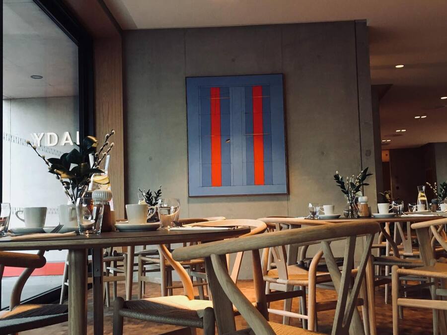 A modern cafe interior with wooden chairs and tables, a large abstract painting on the wall, and a window with a view.