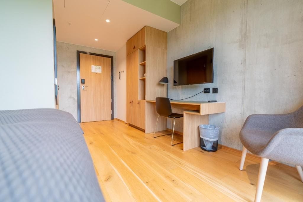 A room with a bed, a desk with a chair, a television, and a trash bin.