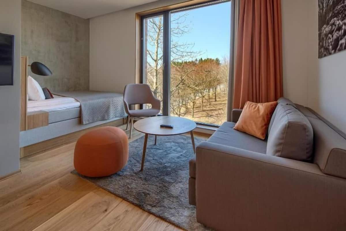A modern living room with a sofa, ottoman, and chair, a small table, and a large window with curtains.