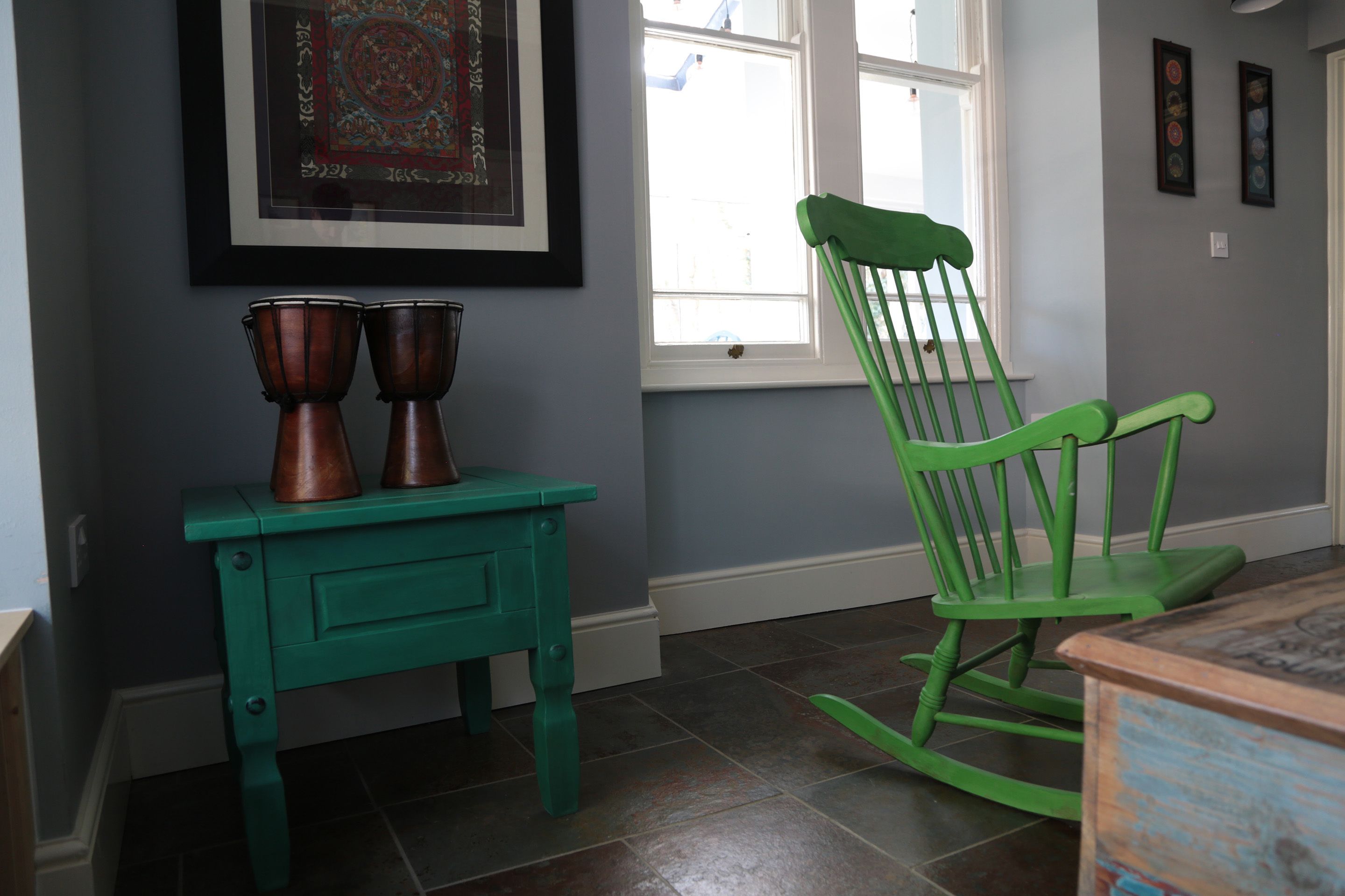 A room with a teal table, two copper pots, a green rocking chair, and a framed artwork on the wall.