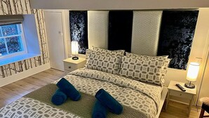 Een slaapkamer met een groot bed, beddengoed met een patroon, een raam met uitzicht en een nachtkastje met een lamp.