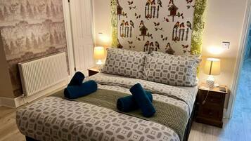 Un dormitorio con una colcha estampada, dos almohadas, un cabecero con motivos de pájaros y hojas, un radiador y lámparas de noche.