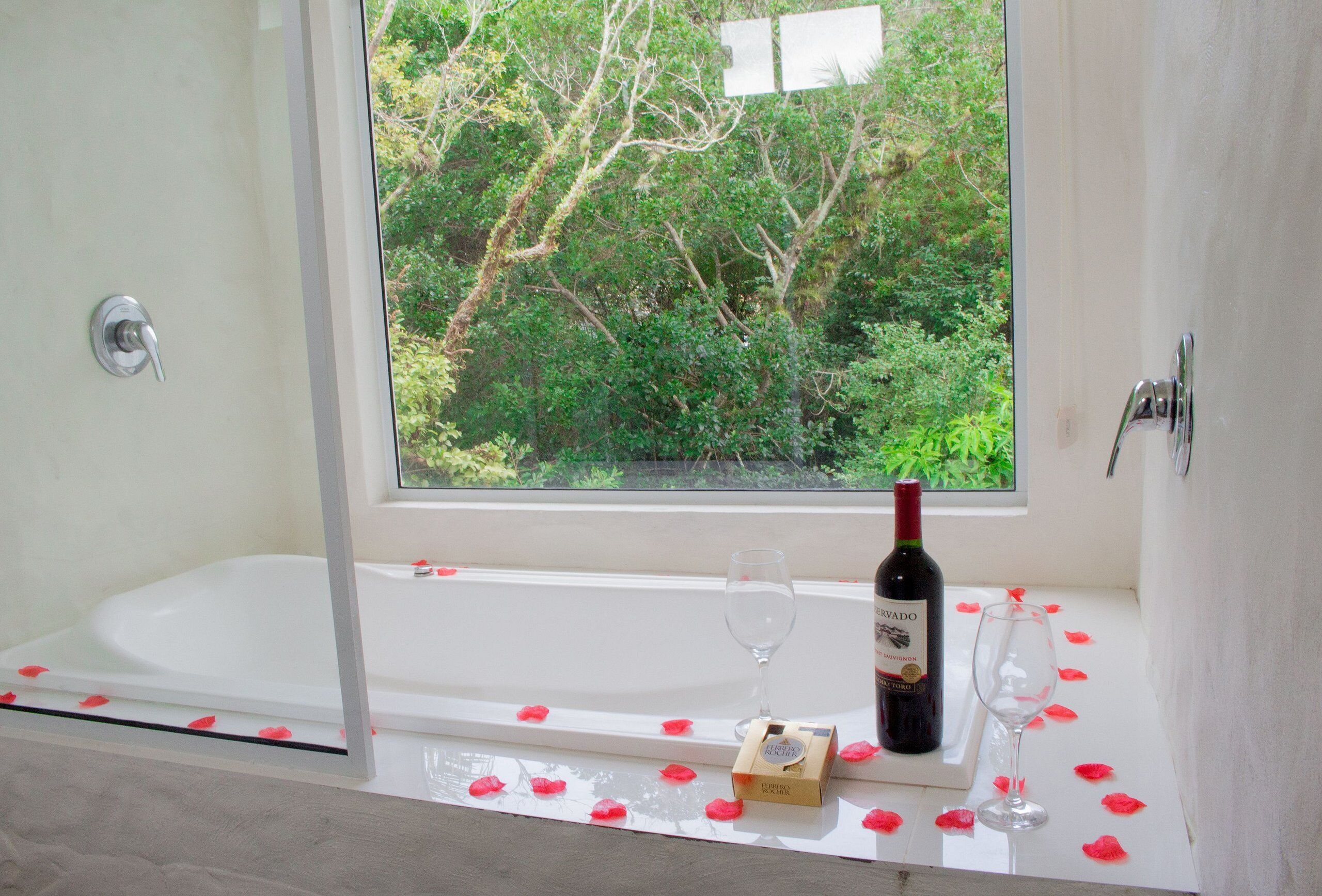 Un bagno con vasca, una finestra vista verde, una bottiglia di vino e un bicchiere sul davanzale.