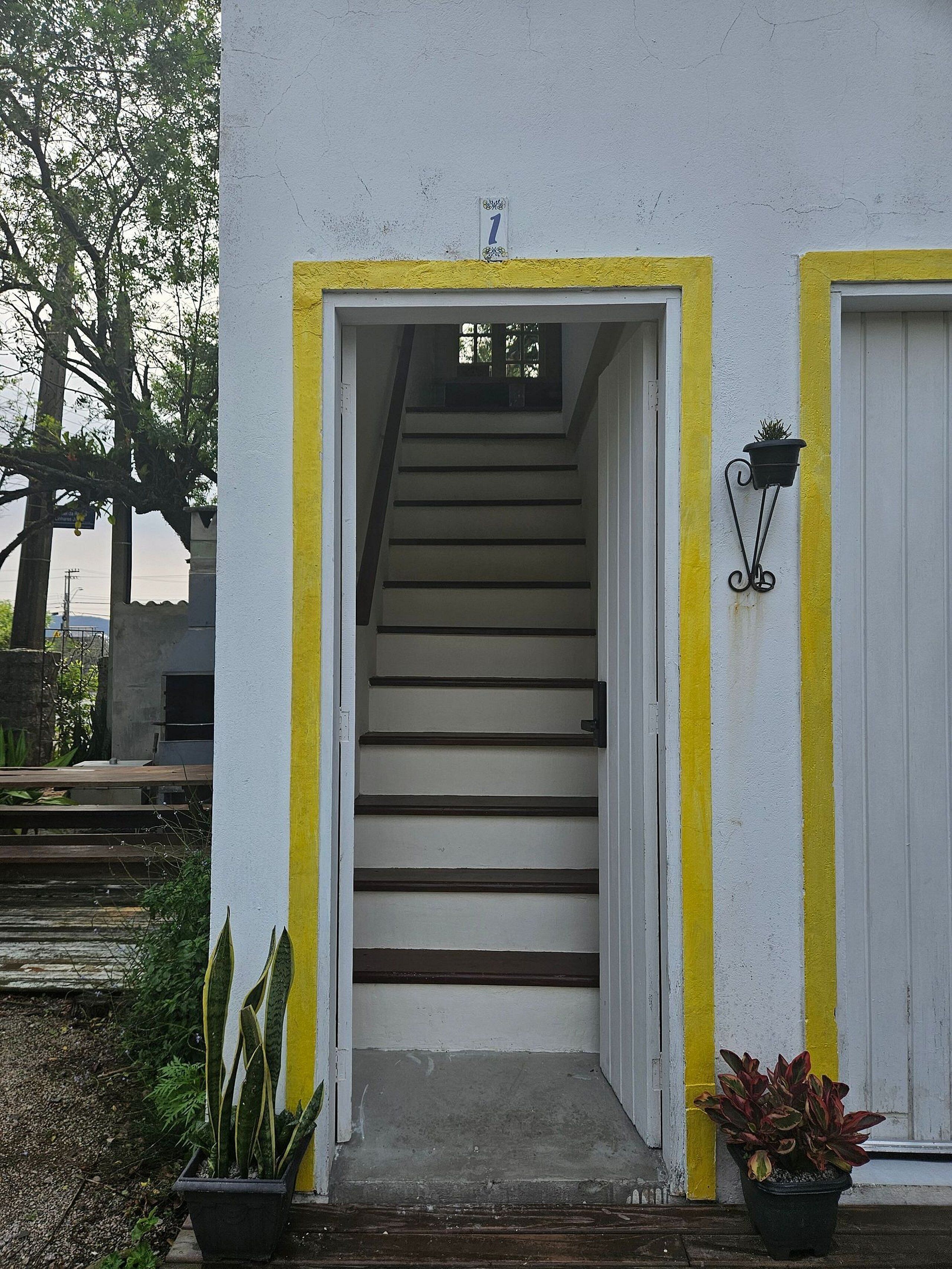 Uma escadaria leva a uma porta com um sinal de número acima, ladeada por paredes pintadas de amarelo e branco, e plantas em vasos na base.