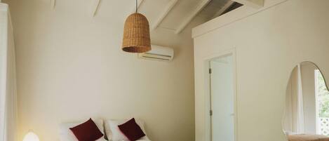 Quarto com cama, duas almofadas, luminária pendente, aparelho de ar condicionado e porta.