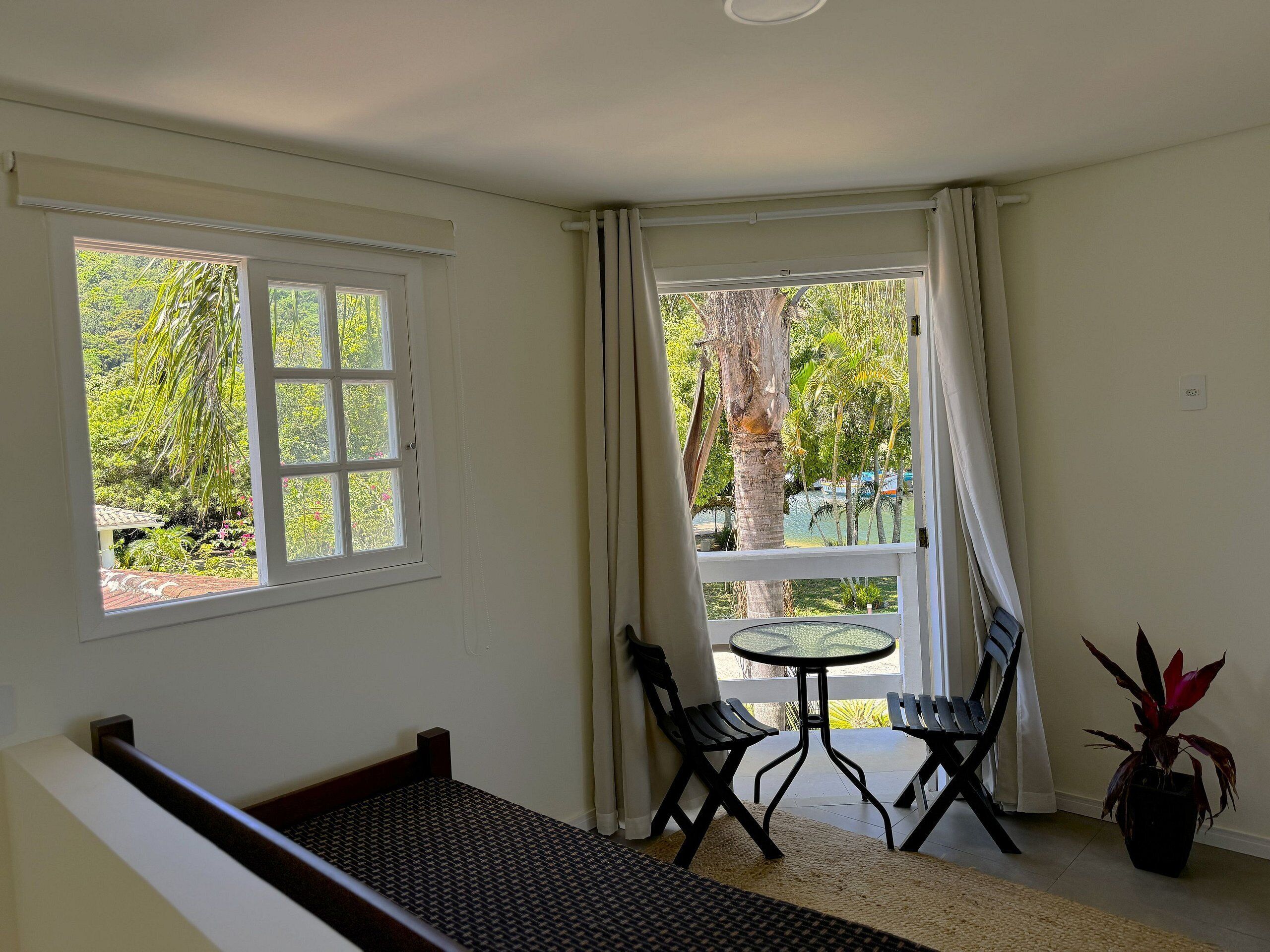 Una habitación con una cama, una mesita con sillas, una planta en maceta y ventanas grandes con cortinas.