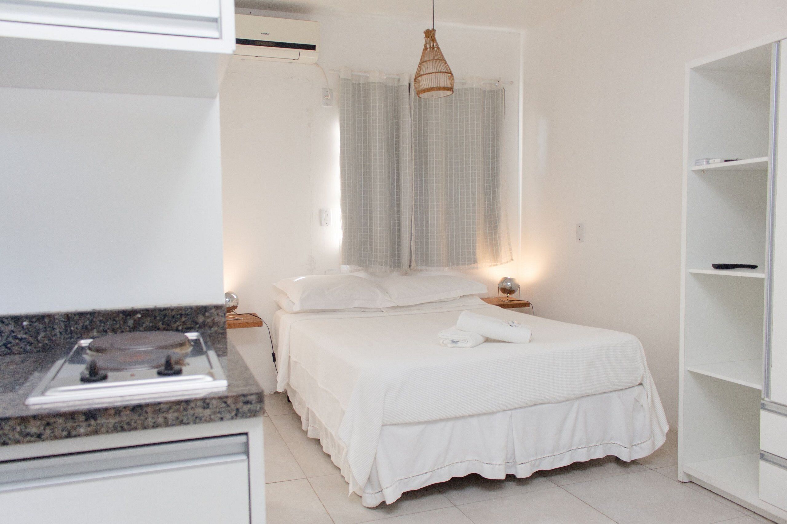 Un dormitorio con cama, mesitas de noche, ventana con cortinas, lámpara colgante y un armario blanco.