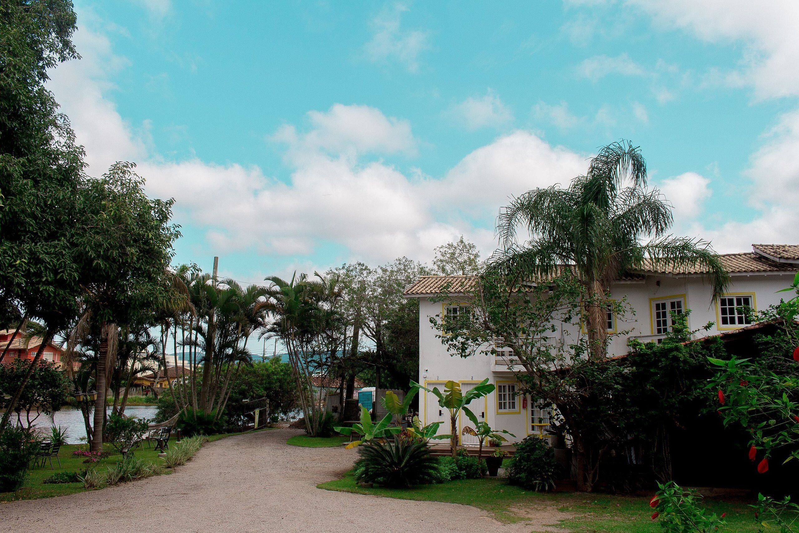 A white building with a tiled roof, surrounded by lush greenery and a gravel path leading to it.