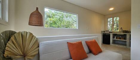 A modern living room with a grey sofa, two orange cushions, a wicker pendant light, and a view of greenery through a window.