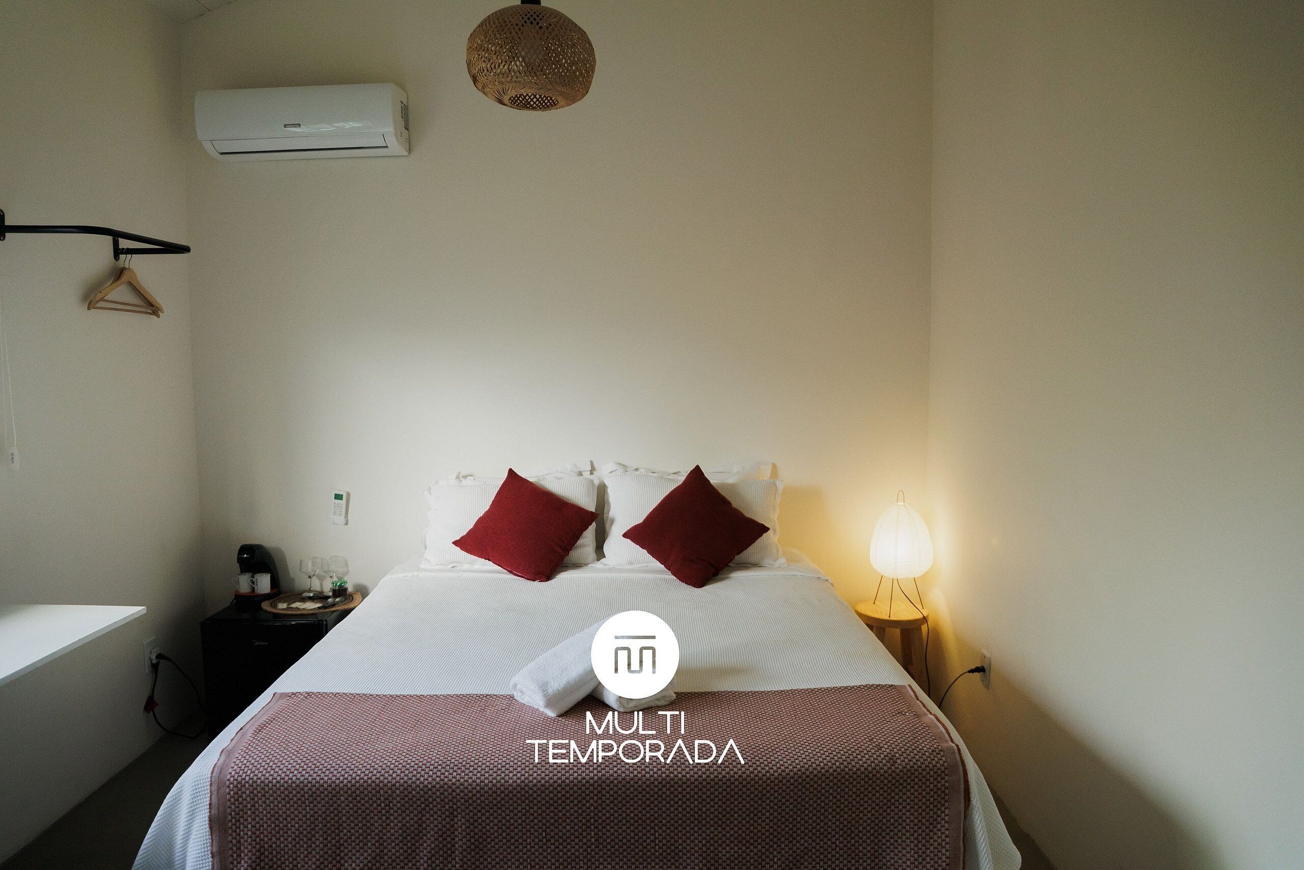 A hotel room with a bed, a bedside table with a lamp, a wall-mounted air conditioner, and a hanging coat rack.