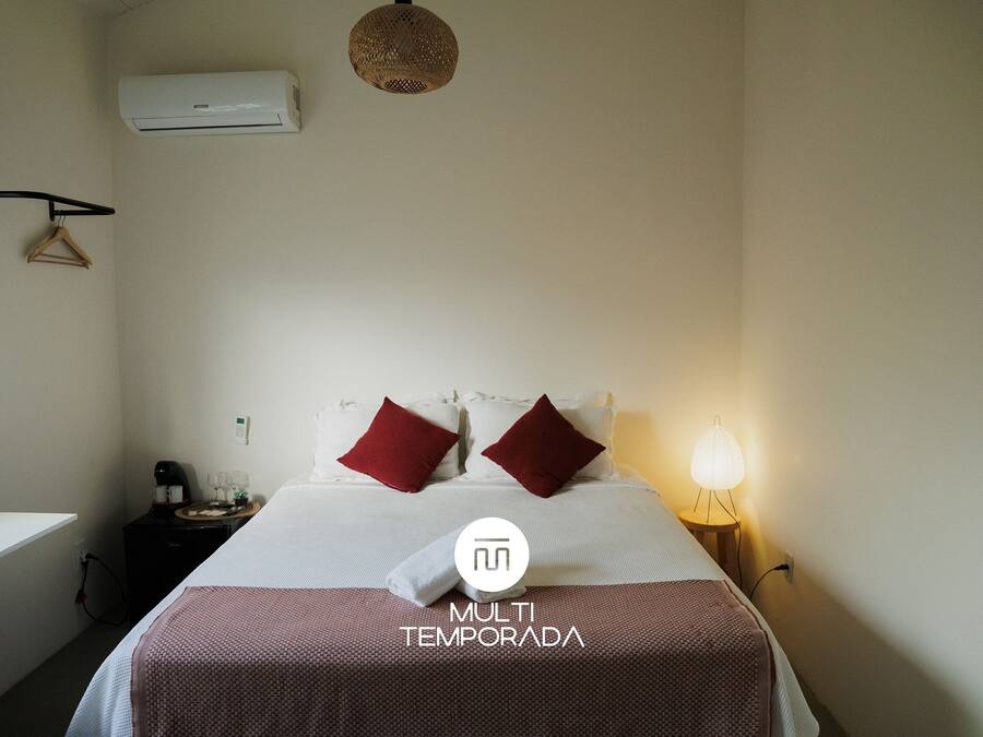 A hotel room with a bed, a bedside table with a lamp, a wall-mounted air conditioner, and a hanging coat rack.