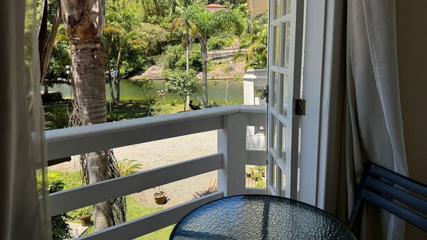 A balcony with a glass table, white railing, and a view of a garden and pond. - Quinta da Margem (Florianópolis)