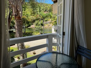 Uma varanda com mesa de vidro, corrimão branco e vista para um jardim e lago.