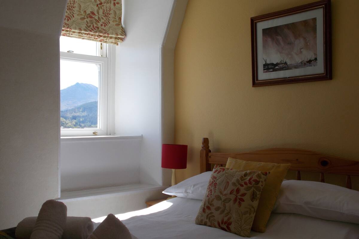 A bedroom with a bed, a window with a view of mountains, a painting on the wall, and a red lamp.