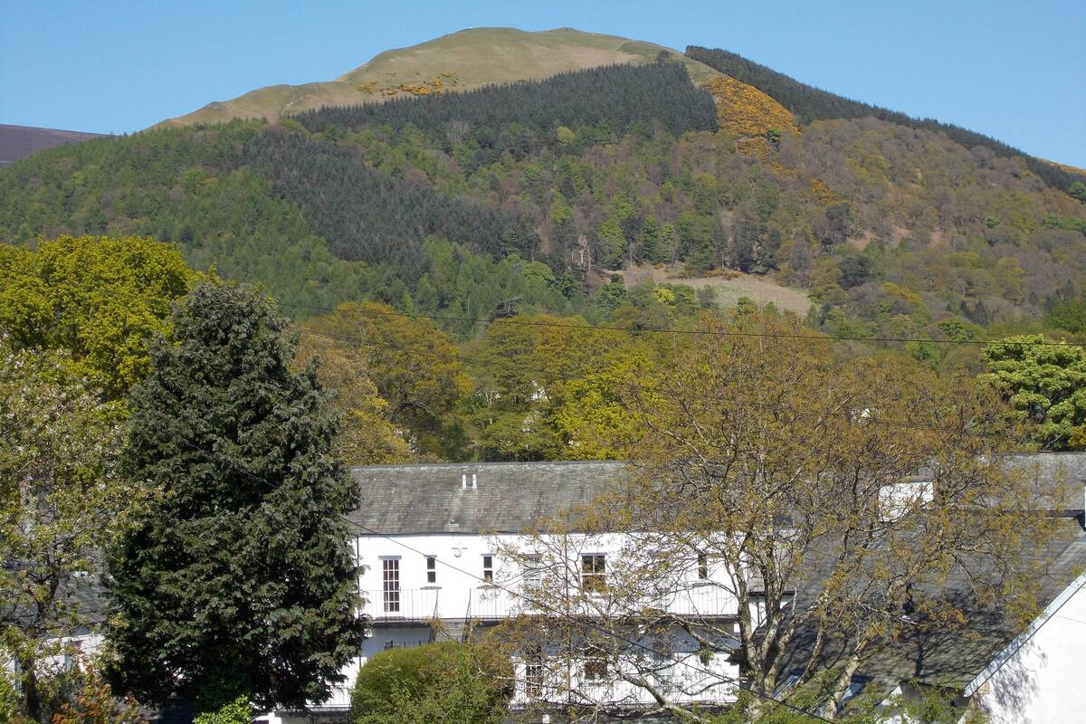 A white building with a grey roof, surrounded by trees and a hill in the background.