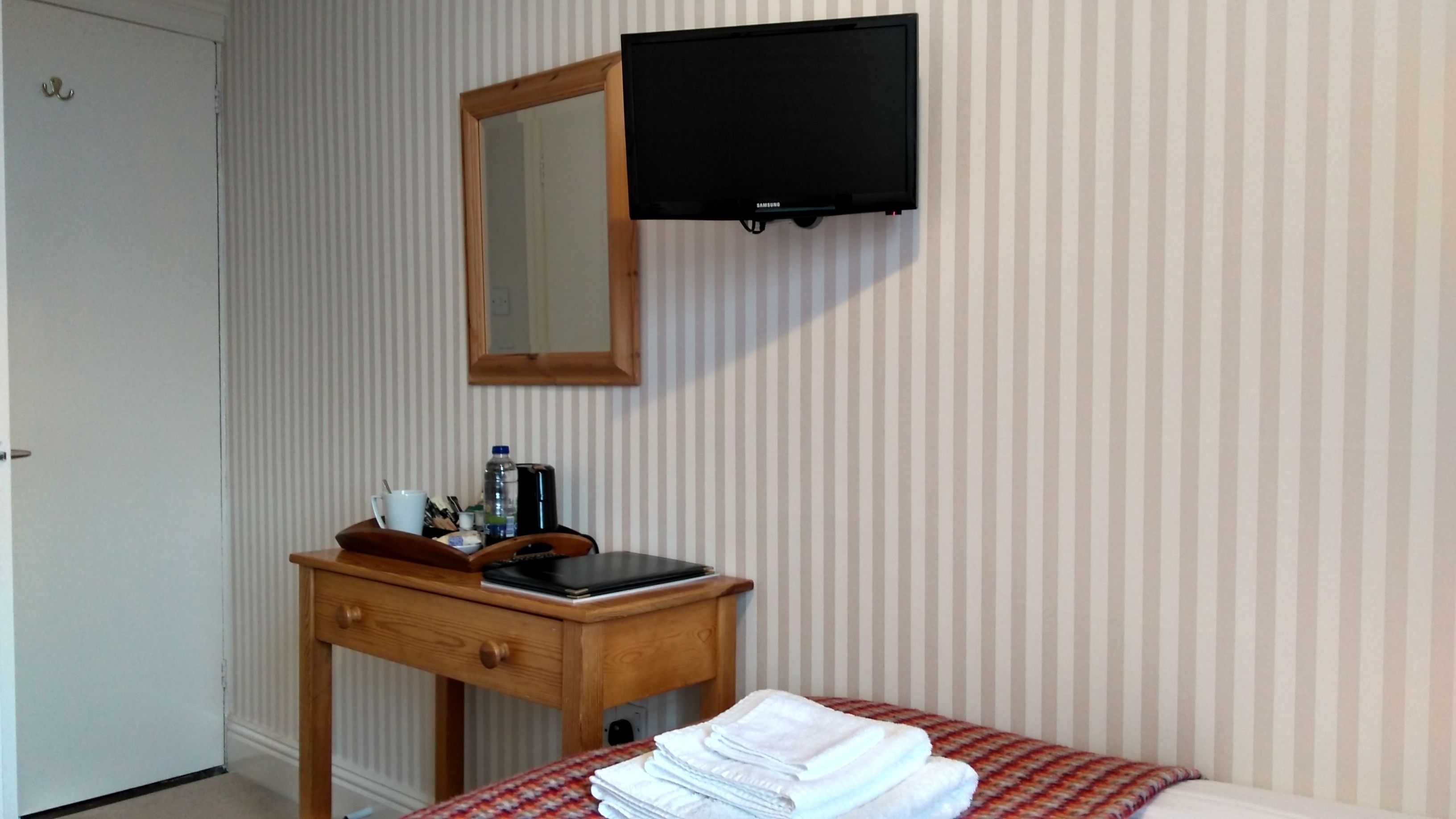 Habitación de hotel con una cama, una mesita de noche con lámpara, un televisor montado en la pared y un espejo sobre la mesita.