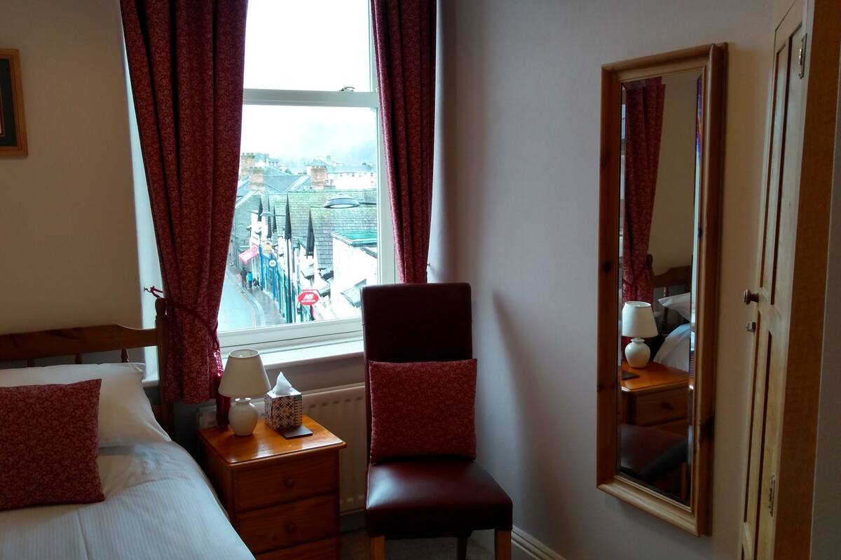 A hotel room with a bed, a chair, a nightstand, a lamp, a mirror, and a window with a view of the city.