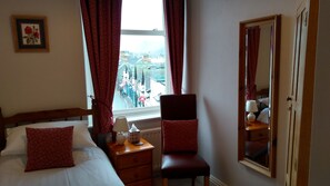 A hotel room with a bed, a chair, a nightstand, a lamp, a mirror, and a window with a view of the city. - Swiss Court (Keswick)