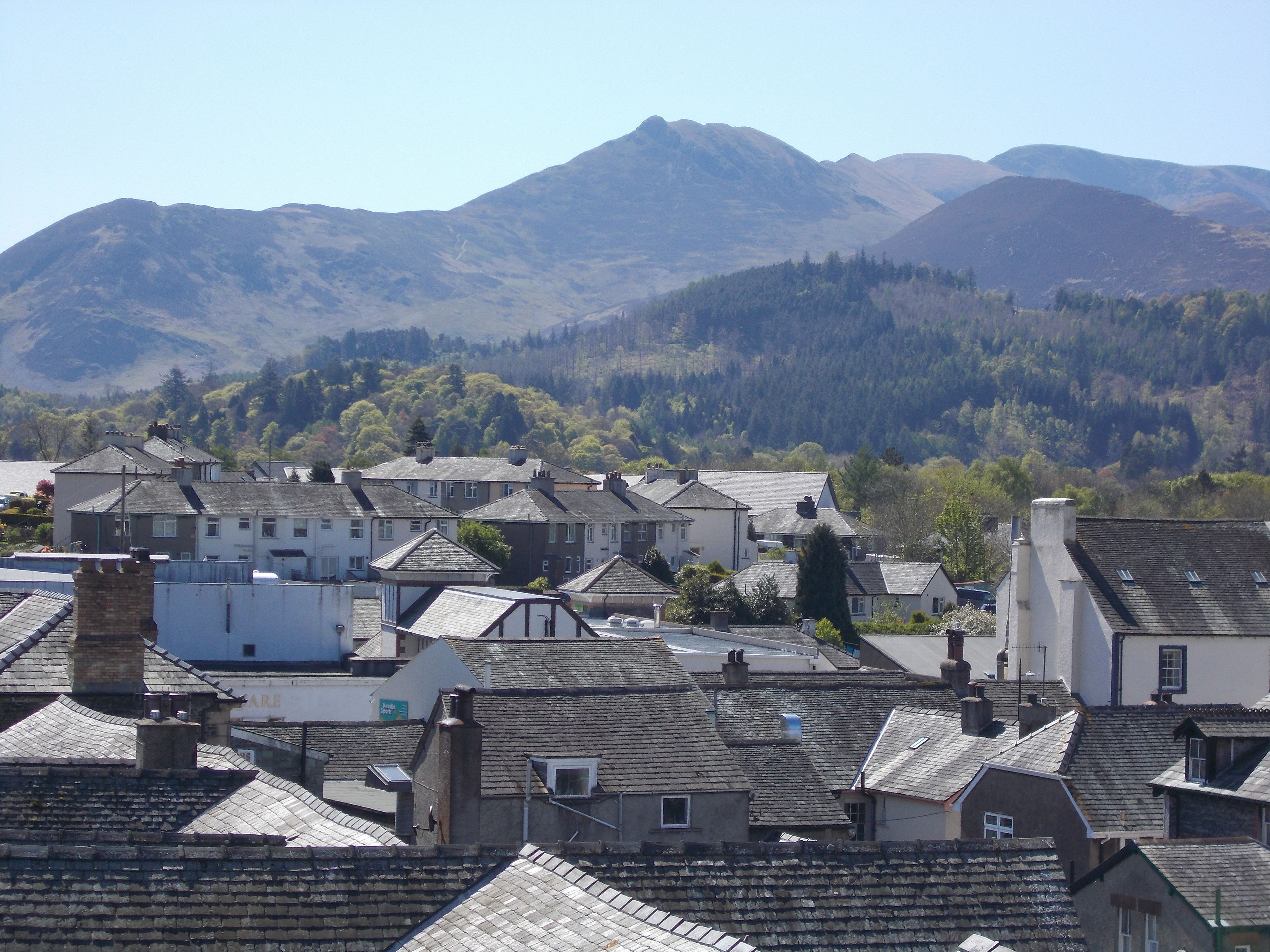A town with traditional buildings, some with visible chimneys and flat roofs, set against a backdrop of mountains and a forested area.