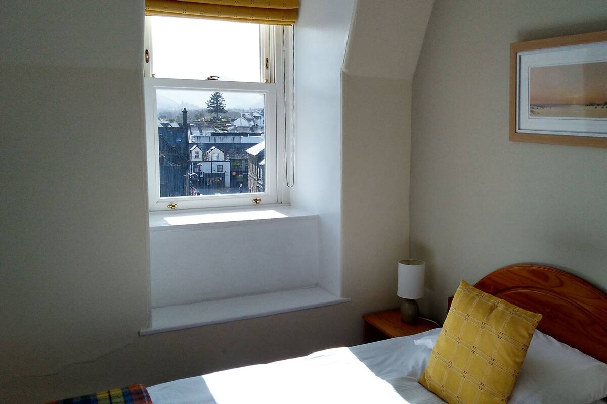 A bedroom with a single bed, a window with a view of buildings, a bedside table with a lamp, and a framed picture on the wall.