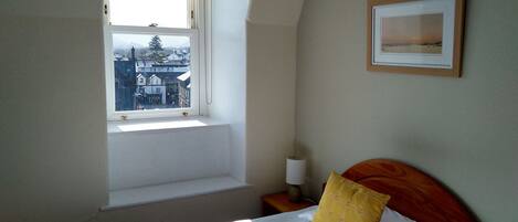 A bedroom with a single bed, a window with a view of buildings, a bedside table with a lamp, and a framed picture on the wall.