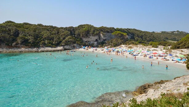 Una playa con agua turquesa cristalina, gente nadando y tomando el sol, y una costa rocosa con árboles.