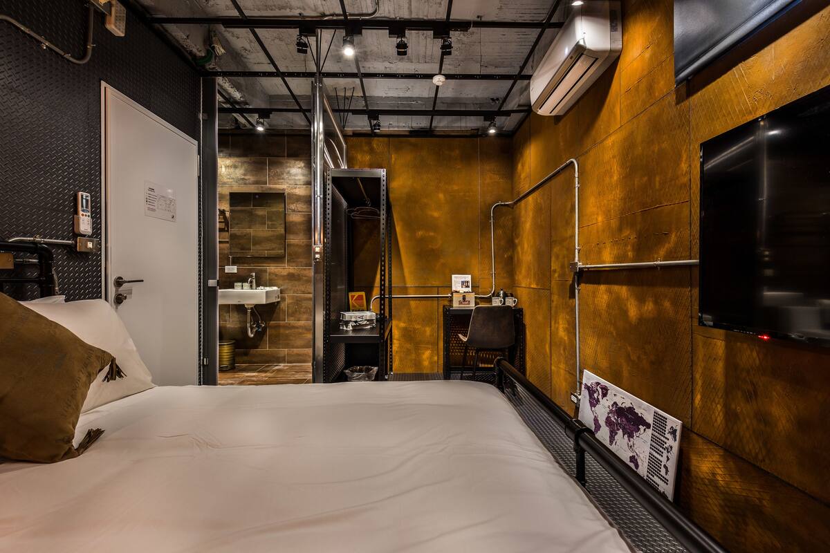 A hotel room with a bed, a desk, a chair, a TV, and a bathroom with a sink and mirror.