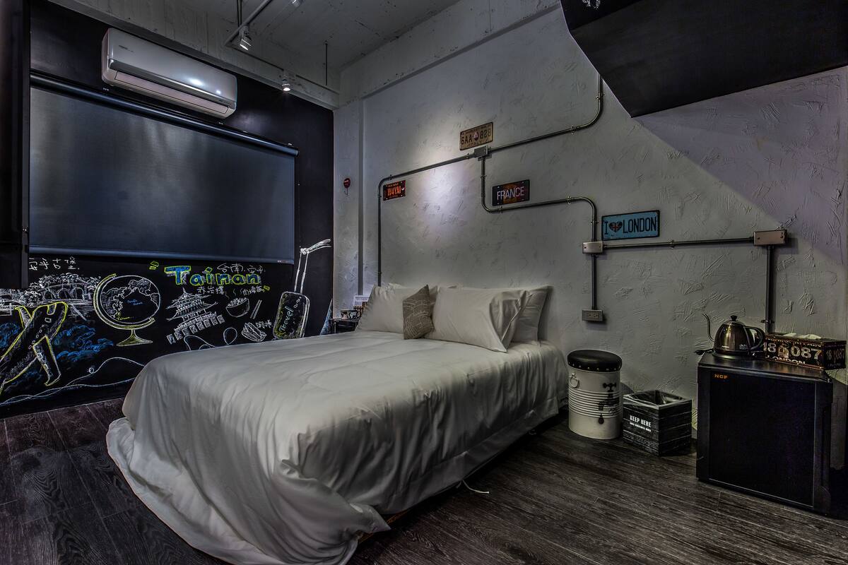 A modern bedroom with a large bed, a small desk, and a wall-mounted TV. There are signs indicating directions to different rooms and a sign that says "I ♥ LONDON".