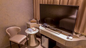 A hotel room with a large flat-screen TV, a small table with a cup and a box of tissues, a chair, and a minibar. - Reality Design Inn (Tainan)