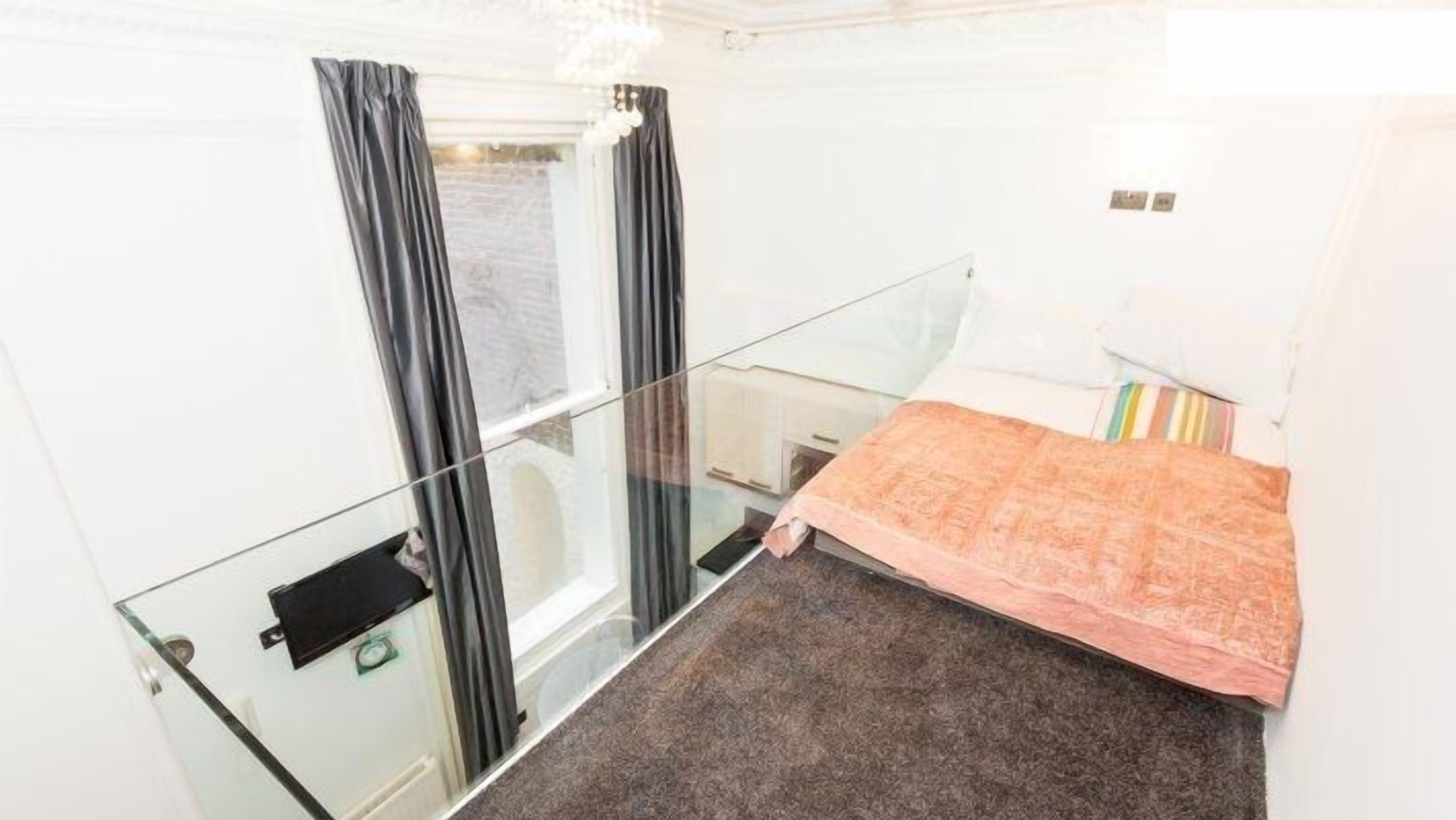 A hotel room with a glass partition, a bed with a pink bedspread, and a window with curtains.