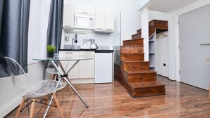 A modern kitchen with white cabinets, a microwave, and a coffee machine. There is a glass table with a clear chair, a wooden staircase, and a wooden floor.