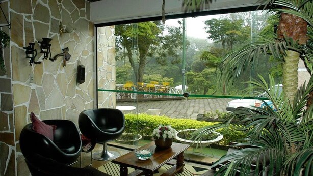 Una habitación con pared de piedra, ventana con vista a un jardín y coches aparcados, dos sillas negras, una mesa de madera con un cuenco de flores, y una pequeña planta.