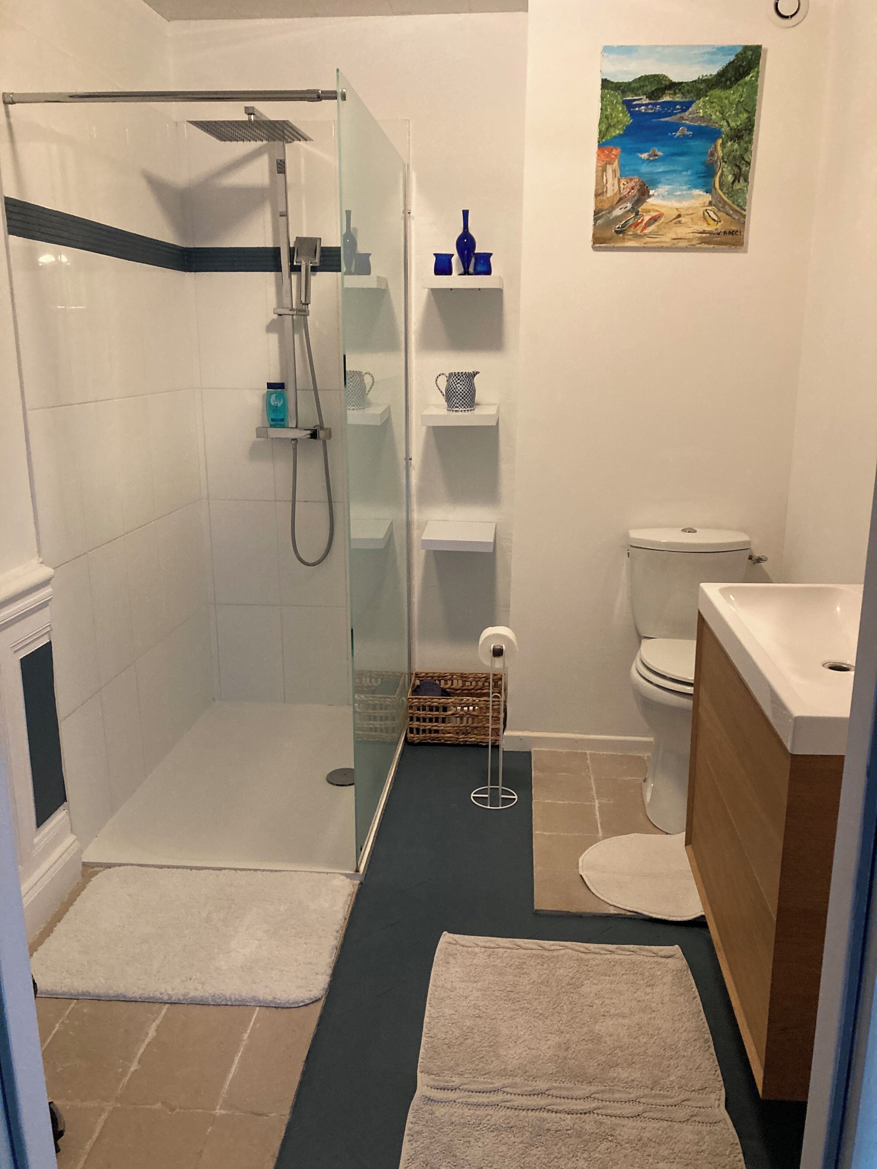 Un baño moderno con cabina de ducha de vidrio, azulejos blancos, un cuadro de una escena costera y un mueble de tocador de madera.