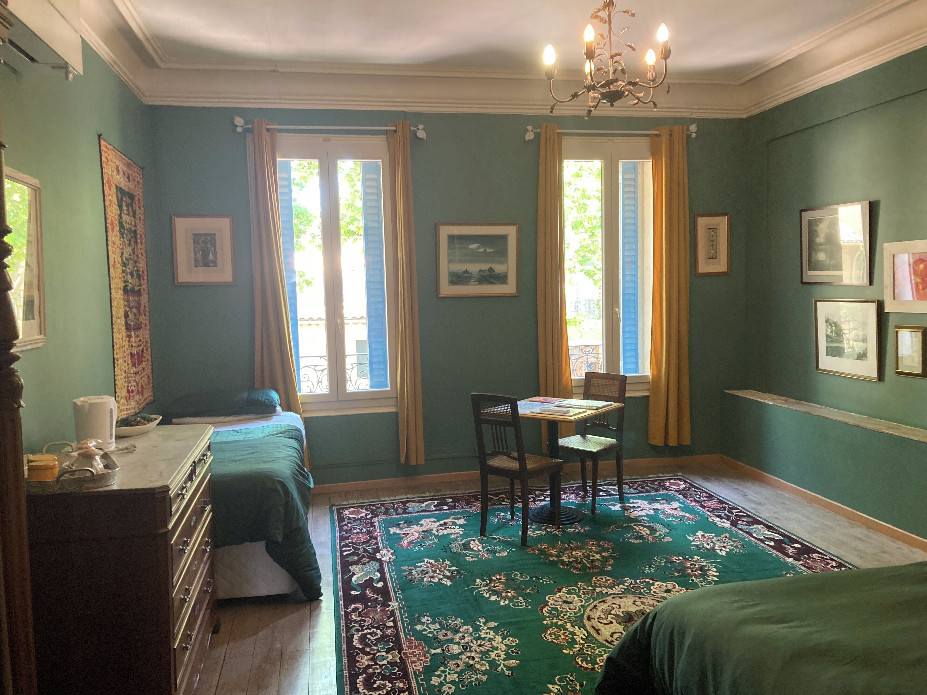 A room with a bed, a desk with a chair, a chandelier, and a large window with curtains.