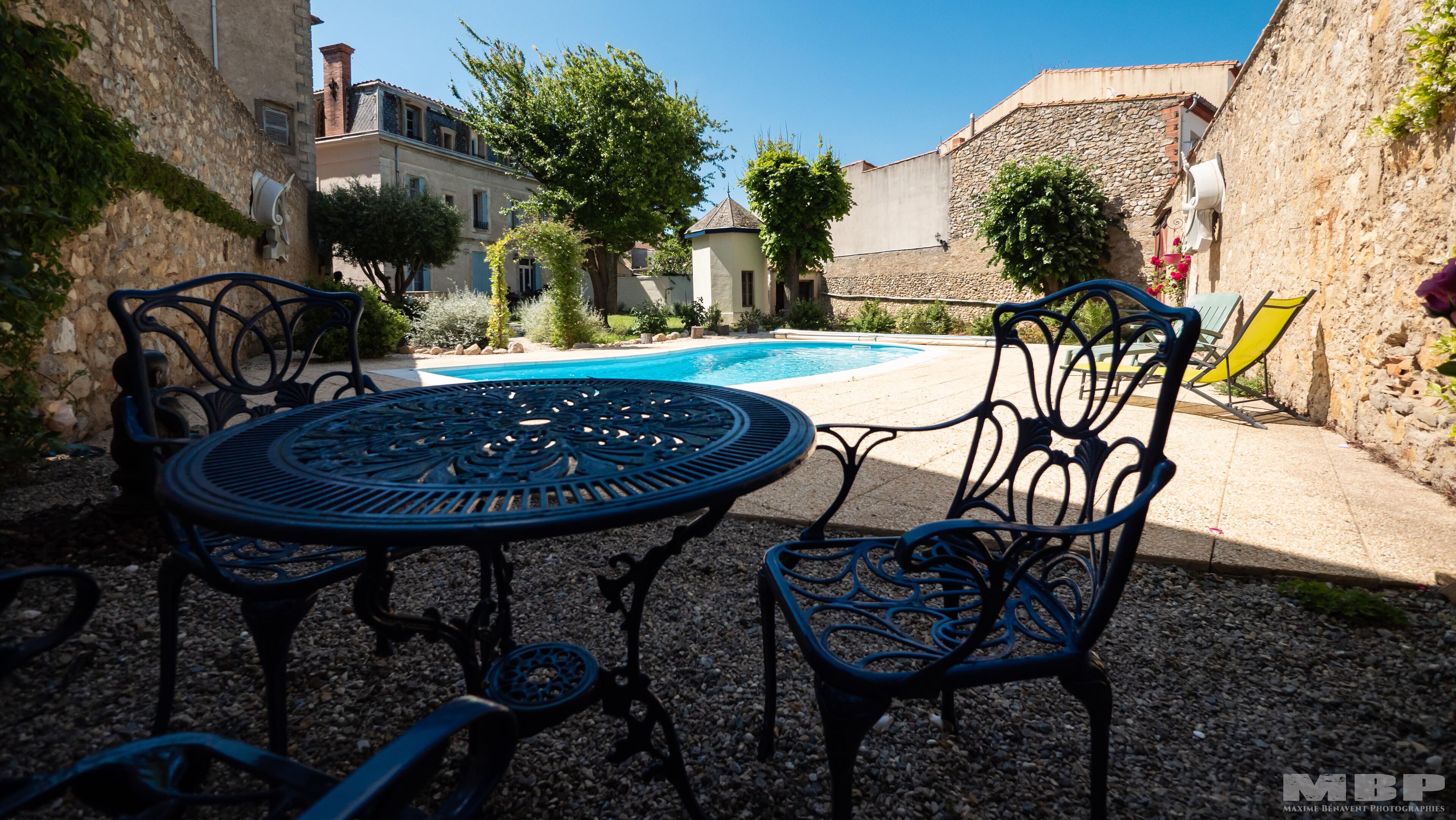 A patio with a round table and chairs, a swimming pool, and stone buildings in the background.