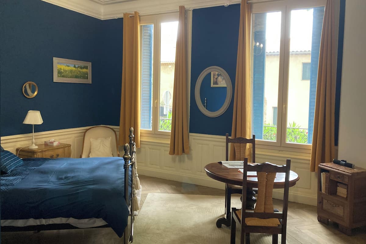 A bedroom with a bed, a round table, and chairs, a window with curtains, and a mirror on the wall.