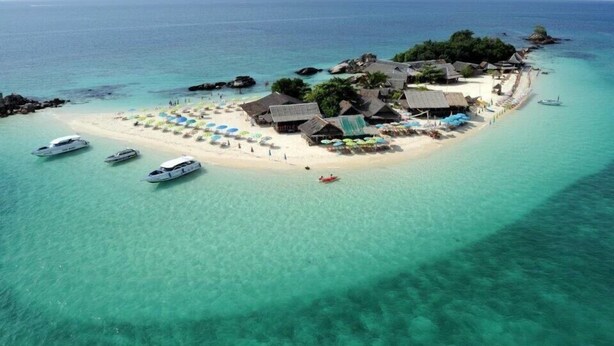 An aerial view of a tropical island with clear turquoise waters, a sandy beach, and several thatched-roof buildings. There are boats docked near the shore and umbrellas set up for sunbathing. - Banhalawee (Ko Yao)