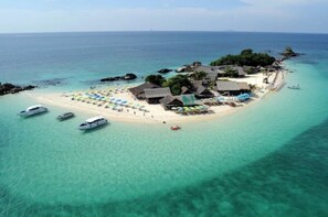Ein Luftbild einer tropischen Insel mit klarem türkisblauem Wasser, einem Sandstrand und mehreren strohgedeckten Gebäuden. Boote liegen an der Küste vor Anker und Sonnenschirme stehen zum Sonnenbaden bereit.