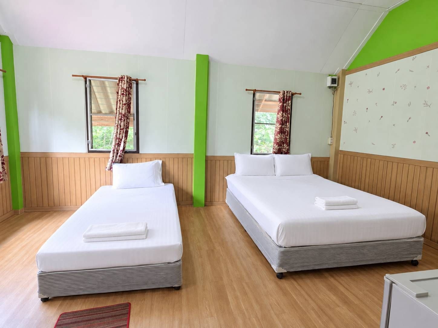A hotel room with two beds, wooden flooring, a green accent wall, and a window with curtains.