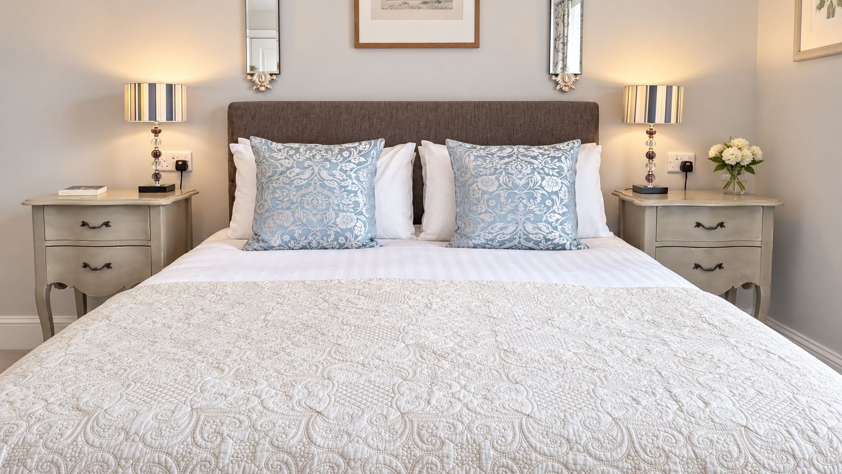 A neatly made bed with a patterned bedspread and two decorative pillows, flanked by bedside tables with lamps and a vase of flowers.