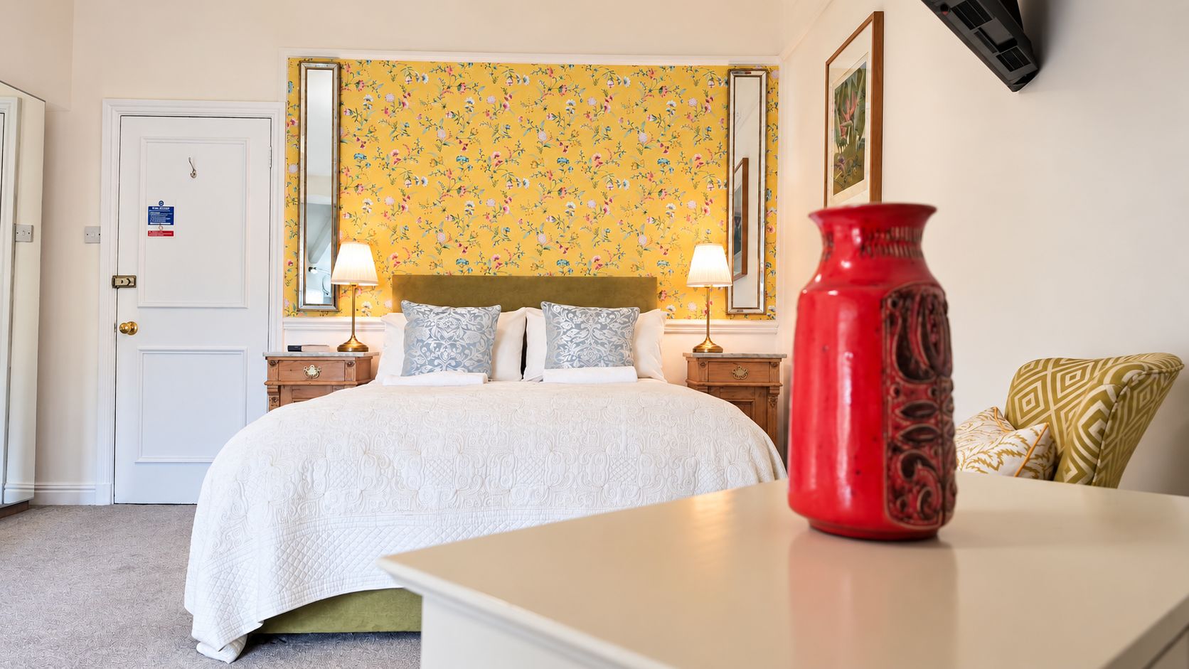 A hotel room with a bed, bedside tables, a red vase, and a floral wallpaper.