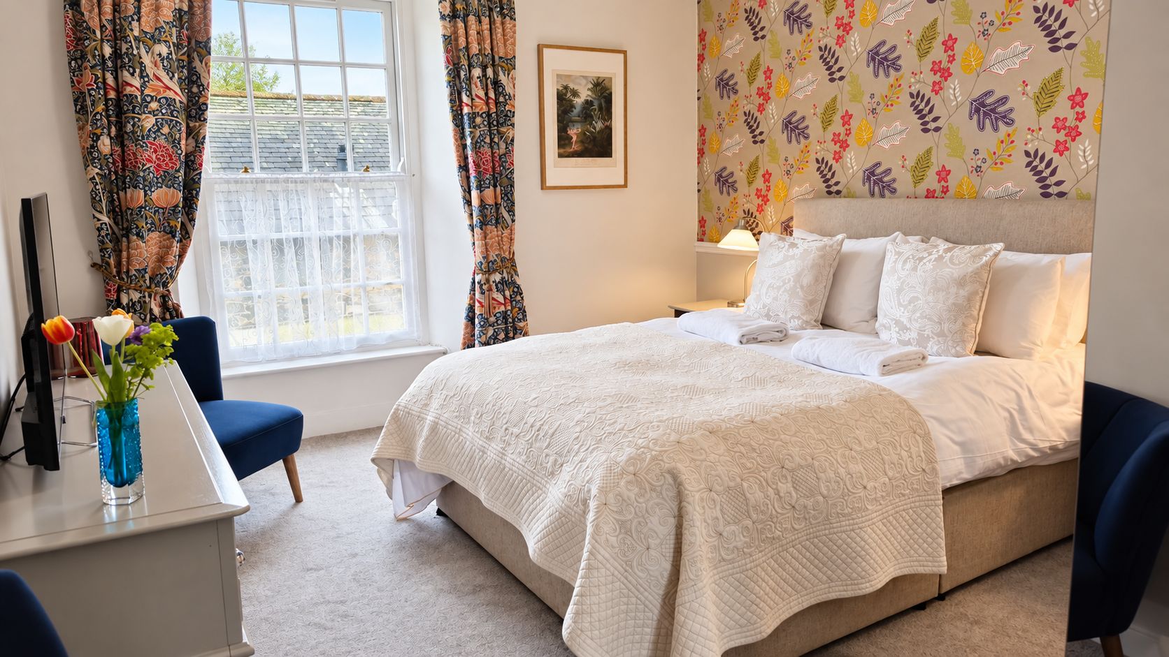 Une chambre à coucher avec un lit, un bureau, une chaise, un vase de fleurs et une fenêtre avec des rideaux.
