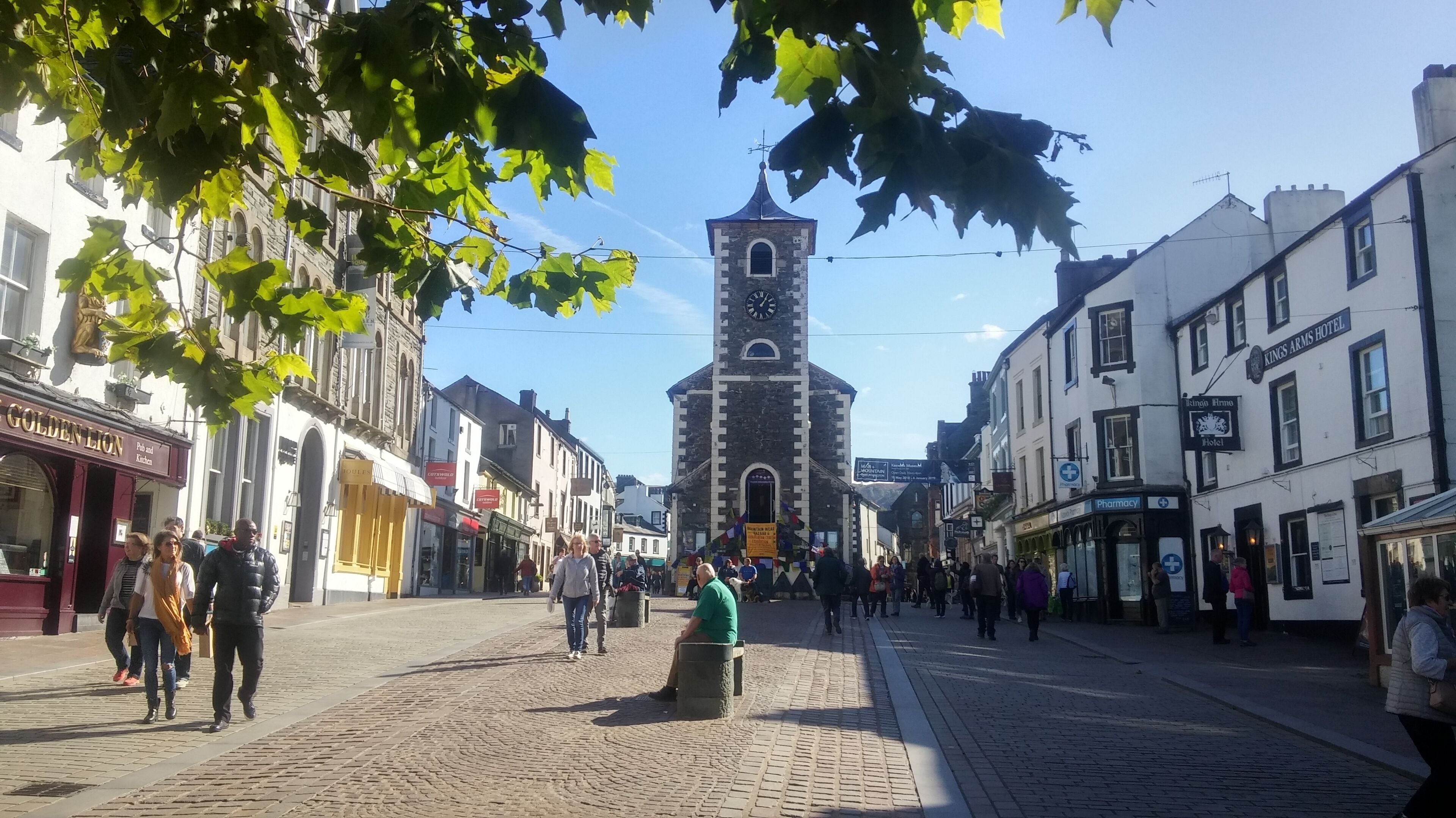 Une place historique bordée de rues pavées, sur laquelle se dressent un imposant beffroi et divers bâtiments commerciaux, notamment un pub et un hôtel.