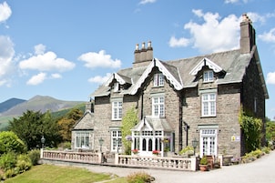 A stone building with multiple windows and a chimney, surrounded by greenery and mountains in the background.