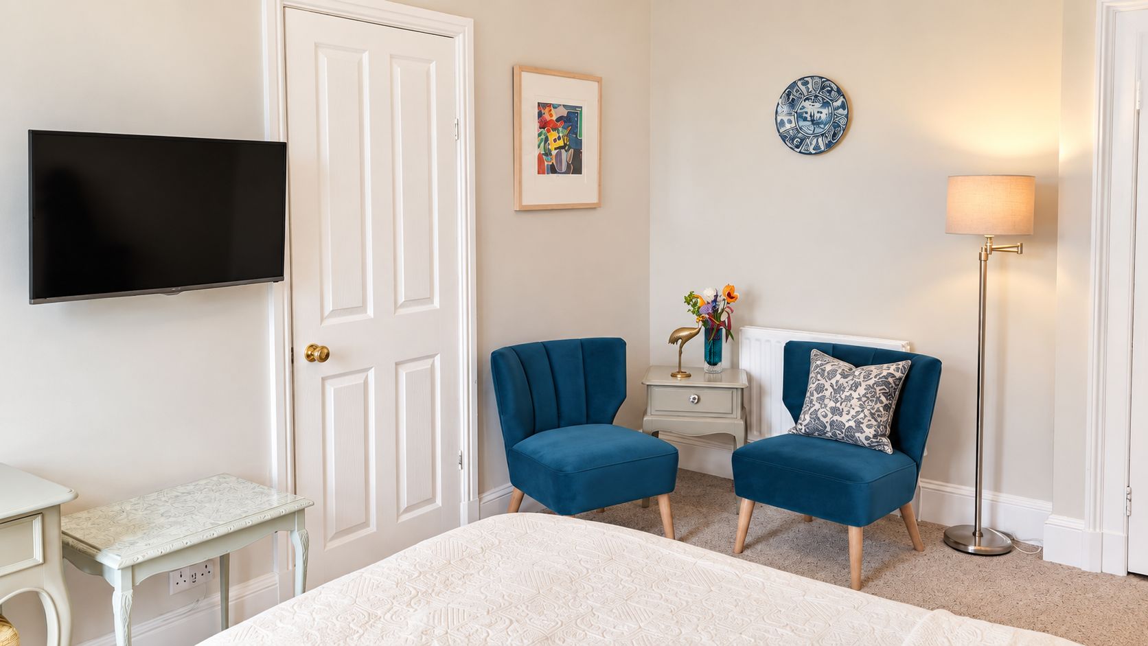 A bedroom with a bed, two blue armchairs, a white bedside table, a TV mounted on the wall, a floor lamp, and a decorative plate.